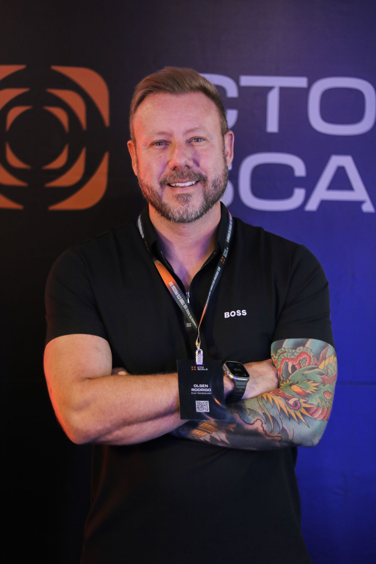 Olsen Rodrigo em pé, de braços cruzados, com crachá no peito em um evento.